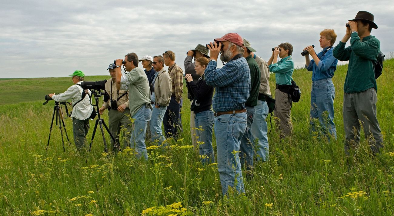 A group of people stand in a grassy field, looking into the distance, some through binoculars.