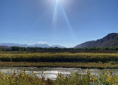 A tranquil river winds through lush greenery beneath a radiant sunbeam, with distant mountains rising under a clear blue sky.
