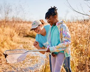 A woman wearing a TNC hat and a young girl smile while observing the contents of a dip net in a field.