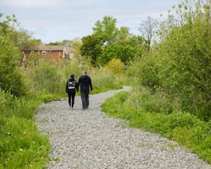 Couple, holding hands, walks on a gravel path surrounded by greenery, with a brick house in the background.